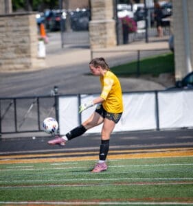 Goalkeeper in a yellow jersey kicking a soccer ball during a game on a sunny day, showcasing athleticism and precision.