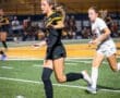 Two female soccer players compete on a field, one dribbling the ball while the other runs closely behind.