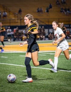 Two female soccer players compete on a field, one dribbling the ball while the other runs closely behind.