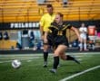 High school girl soccer player #12 kicking a ball during a game, with referee and spectators in the background.