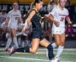 High school girl soccer player in black uniform skillfully dribbles the ball on field during a night game.