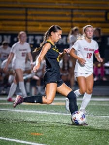 High school girl soccer player in black uniform skillfully dribbles the ball on field during a night game.