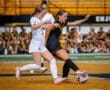Two female soccer players compete for the ball on the field as teammates watch from the sidelines in the background.