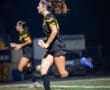 A female soccer player in black and yellow uniform runs on a field under night lights, showcasing athletic action and teamwork.