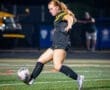 Young female soccer player in action, kicking a ball on a well-lit field during a nighttime game.