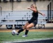 Soccer player in black uniform kicking a ball on a field with bleachers in the background. Action shot on a sunny day.