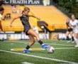 Female soccer player wearing black jersey and blue cleats kicking a ball during a game on a sunny day.