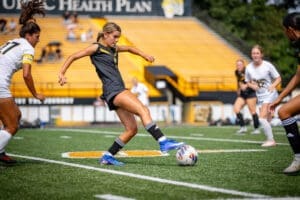 Female soccer player wearing black jersey and blue cleats kicking a ball during a game on a sunny day.