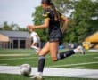 Female soccer player in black uniform dribbles ball on field during a match, with teammate in white in the background.