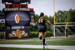 Female soccer player in black jersey dribbles ball on Wright Field, game scoreboard shows time remaining.