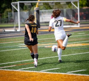 Two female soccer players chase the ball on the field, one in black with “Tigers” and the other in white.