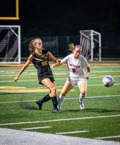 Two female soccer players compete for the ball on a well-lit field at night, showcasing athleticism and teamwork.