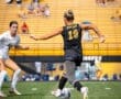 Two female soccer players competing on a field, one dribbling a ball while the other defends, with yellow bleachers in the background.
