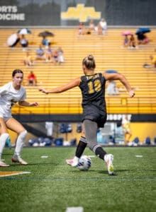 Two female soccer players competing on a field, one dribbling a ball while the other defends, with yellow bleachers in the background.