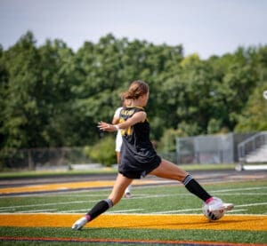 Young female soccer player kicking a soccer ball on a field during a game, showcasing athleticism and energy.
