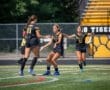 Three female soccer players celebrate a goal on a green turf field, showcasing teamwork and athleticism.