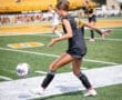 Soccer player in black uniform kicks a ball on the field, showcasing athleticism during an outdoor game.