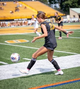 Soccer player in black uniform kicks a ball on the field, showcasing athleticism during an outdoor game.