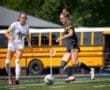 Two female soccer players compete for the ball on a sunny field, with a school bus in the background.