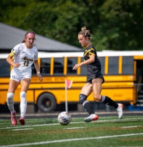 Two female soccer players compete for the ball on a sunny field, with a school bus in the background.
