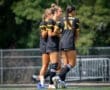 Three female soccer players in black jerseys share a light moment on the field, showcasing teamwork and camaraderie.