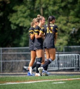 Three female soccer players in black jerseys share a light moment on the field, showcasing teamwork and camaraderie.