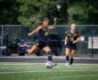 Soccer player in black uniform kicks the ball on a field, with a teammate in the background, showcasing athleticism and teamwork.