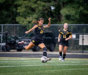 Soccer player in black uniform kicks the ball on a field, with a teammate in the background, showcasing athleticism and teamwork.