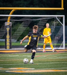 Soccer player in a black jersey kicks the ball on a field, with a goalkeeper in yellow preparing to defend.