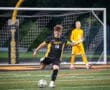 Player in black jersey (18) kicks soccer ball on field, while goalie in yellow stands ready in the background.