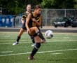 Female soccer player in black uniform skillfully kicking a soccer ball on a field during an outdoor game.
