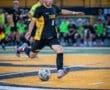 Young soccer player in black and yellow uniform dribbles the ball on the field during a match under stadium lights.