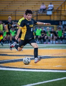 Young soccer player in black and yellow uniform dribbles the ball on the field during a match under stadium lights.