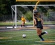 Soccer player wearing black jersey with number 22 runs toward the ball on the field, with a goalie in the background.
