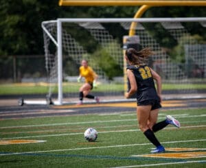 Soccer player wearing black jersey with number 22 runs toward the ball on the field, with a goalie in the background.
