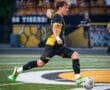 A high school soccer player in action, kicking a ball on the field, with fans cheering in the background.