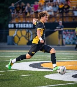 A high school soccer player in action, kicking a ball on the field, with fans cheering in the background.