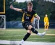 A soccer player in a black jersey kicks the ball on a field, with teammates in the background during an evening game.