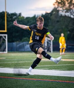 A soccer player in a black jersey kicks the ball on a field, with teammates in the background during an evening game.