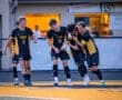 High school soccer players celebrating a goal, wearing black and gold uniforms, on a vibrant field under evening light.