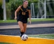 A female soccer player in a black uniform dribbles a ball on a vibrant turf field during a game.