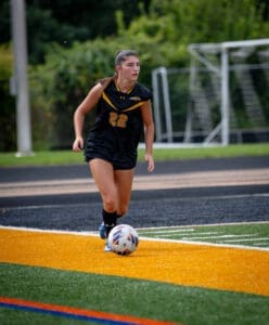 A female soccer player in a black uniform dribbles a ball on a vibrant turf field during a game.
