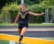 Young female soccer player in black uniform kicking a soccer ball on a vibrant field during a game.