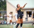 A female soccer player in a black jersey leaps to head the ball during a game, with teammates in white behind her.