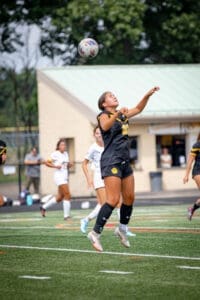 A female soccer player in a black jersey leaps to head the ball during a game, with teammates in white behind her.