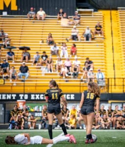 Two female soccer players stand on the field, while a teammate lies on the ground, with spectators in the background.