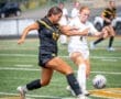 Two female soccer players, one in black and one in white, compete for the ball on a grass field during a match.