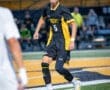Young soccer player in black and yellow uniform dribbling a ball on the field during a night match.