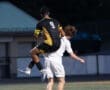 Soccer players competing for the ball, one leaping over the other in an intense match on the field.