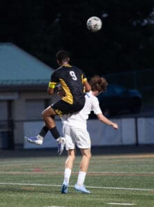 Soccer players competing for the ball, one leaping over the other in an intense match on the field.